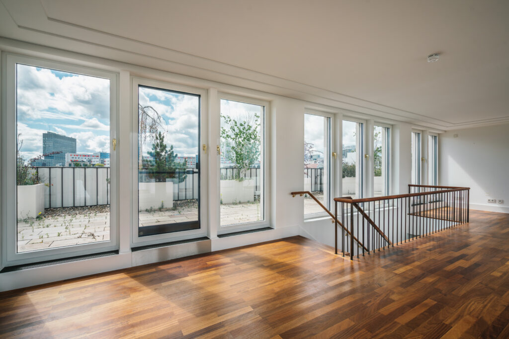 Modernes, lichtdurchflutetes Penthouse mit bodentiefen Fenstern und einer großen Terrasse. Der Raum hat einen edlen Holzfußboden und eine stilvolle Treppe mit Metallgeländer, die in eine untere Etage führt. Durch die Fensterfront bietet sich ein beeindruckender Blick auf eine städtische Skyline mit modernen Gebäuden.