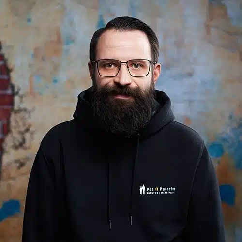 Porträt eines Mannes mit dunklem Vollbart, Brille und schwarzem Hoodie mit dem Logo von „Pat & Patachon“. Er steht vor einer künstlerisch gestalteten Wand mit einem gemalten Schattenriss einer Person aus Backstein und lächelt dezent in die Kamera.