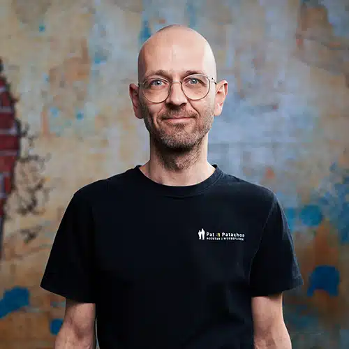 Porträtbild eines lächelnden Mannes mit Glatze. Er trägt eine Brille und ein schwarzes T-Shirt mit Pat & Patachon Logoaufdruck. Er steht vor einer künstlerisch gestalteten Wand mit einer Pat & Patachon Backstein-Silhouette.
