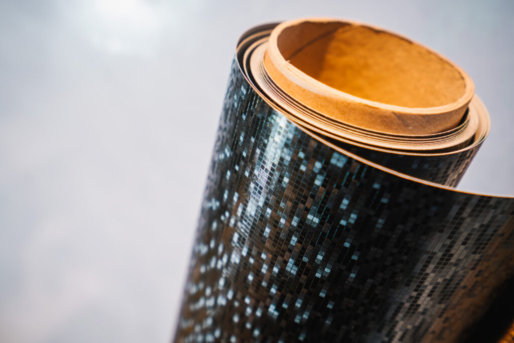 Eine Rolle hochwertiger Möbelfolie mit einer glänzenden, pixelartigen schwarzen Struktur. Die Folie reflektiert das Licht in unterschiedlichen Winkeln und erzeugt eine moderne, dynamische Optik. Der Kartonkern der Rolle ist sichtbar, während sich die Folie sanft aufrollt. Der Hintergrund ist weich und unscharf, wodurch die glänzende Textur der Folie im Fokus steht.