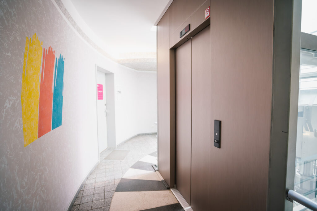Ein moderner Aufzug mit einer hochwertigen, metallischen Folienbeschichtung an den Türen und der Wandverkleidung. Der Fahrstuhl befindet sich in einem hellen Flurbereich mit geschwungener Architektur. An der Wand links ist eine künstlerische Farbgestaltung mit drei Pinselstrichen in Gelb, Orange und Blau zu sehen. Die Glasfront rechts lässt natürliches Licht in den Raum und sorgt für eine freundliche Atmosphäre.