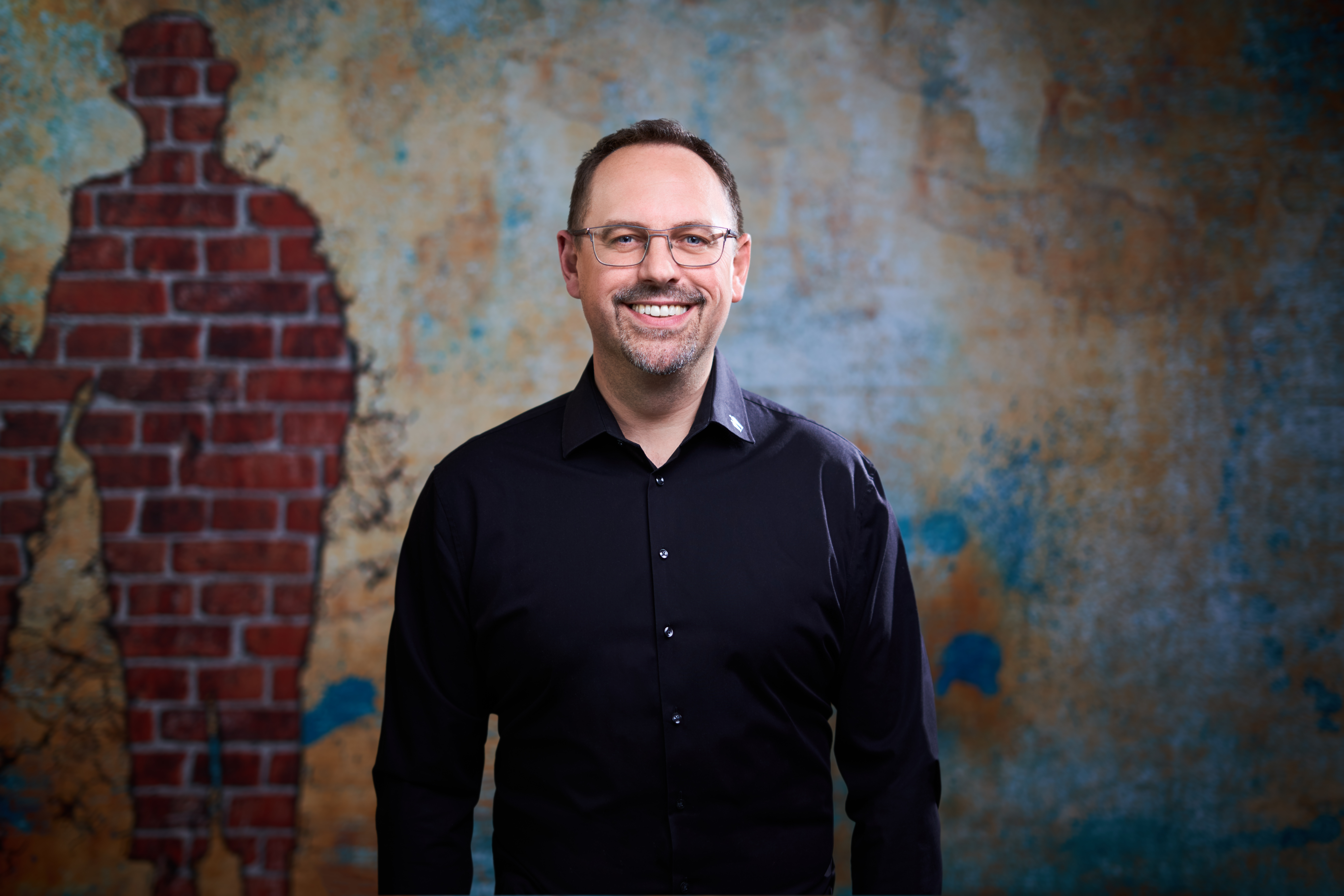 Porträt eines lächelnden Mannes mit Brille, kurzem dunklem Haar und leichtem Bart. Er trägt ein schwarzes Hemd mit einer dezenten Pat & Patachon Logo Stickerei am Kragen. Der Hintergrund ist eine künstlerische Wand mit einer rustikalen, verwitterten Textur in Blau- und Brauntönen. Links im Hintergrund ist eine stilisierte menschliche Silhouette aus Backsteinmustern in die Wand integriert.