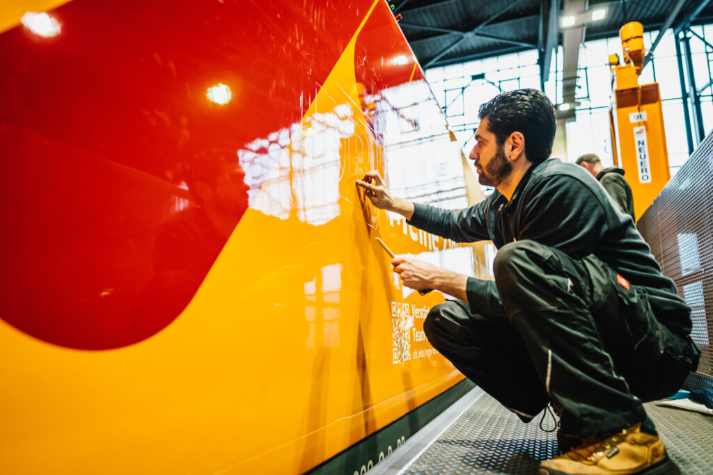 Ein Monteur kniet vor einer glänzend folierten Bahn in Rot und Gelb und arbeitet präzise an einer Beschriftung. Mit einem Werkzeug in der Hand bringt er Details auf der Oberfläche an. Die reflektierende Oberfläche des Fahrzeugs spiegelt das Licht der Umgebung und den Arbeiter selbst. Im Hintergrund sind eine moderne Werkstatthalle mit großen Fenstern und weitere Arbeiter zu sehen
