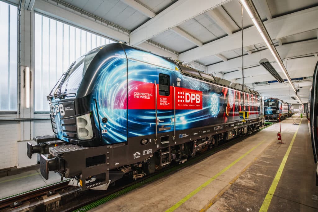Eine moderne Elektrolokomotive des Typs Siemens Vectron steht in einer Wartungshalle. Die Lokomotive ist mit einer auffälligen blauen Folierung gestaltet, die ein dynamisches Lichtdesign zeigt, kombiniert mit dem Logo 'DPB Rail Infra Service GmbH'. Ein rotes Banner mit der Aufschrift 'Connecting the World' ist ebenfalls auf der Seite angebracht. Die Lok steht auf einem Gleis mit grünen Markierungen, umgeben von industriellen Strukturen wie Rohren, Kabeln und Feuerlöschern an der Wand. Die Perspektive des Bildes führt entlang der Lokomotive in die Tiefe der Halle.