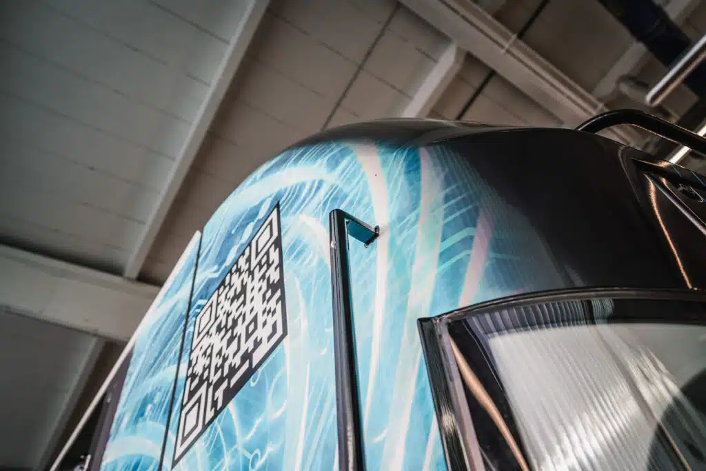 Nahaufnahme der Frontpartie einer modernen Elektrolokomotive mit einer blauen, digitalen Folierung. Die Gestaltung zeigt ein leuchtendes Netzwerk-Design, das den technologischen Fortschritt symbolisiert. Auffällig ist ein großer QR-Code auf der Seitenfläche der Lok. Im unteren rechten Bereich ist ein Teil des Scheinwerfers zu sehen, während sich im Hintergrund die helle Decke der Wartungshalle mit Stahlträgern erstreckt.