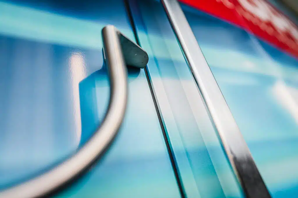 Nahaufnahme eines metallenen Handlaufs und einer Türverkleidung an einem Zug. Die Oberfläche ist mit einer glänzenden blauen Folierung versehen, die Reflexionen des Lichts erzeugt. Ein rotes Designelement ist im oberen Bereich des Bildes sichtbar. Der Fokus liegt auf den Details der Verarbeitung und der hochwertigen Materialstruktur.
