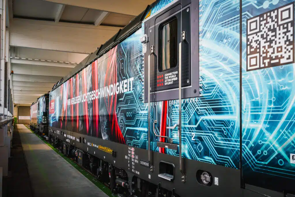 Seitenansicht einer modern folierten Siemens Vectron-Lokomotive in einer Wartungshalle. Die auffällige Gestaltung zeigt ein digitales Leiterbahn-Design in Blau mit roten Lichtstrahlen und trägt die Botschaft 'WIR VERLEGEN LICHTGESCHWINDIGKEIT'. Ein QR-Code ist auf der Folierung zu sehen, ebenso wie der Slogan 'CONNECTING THE WORLD AT THE SPEED OF LIGHT' neben der Einstiegstür. Die Lok steht auf einem Gleis mit grünen Markierungen, und die Halle ist durch ihre weißen Stahlträger und das einfallende Licht erkennbar.