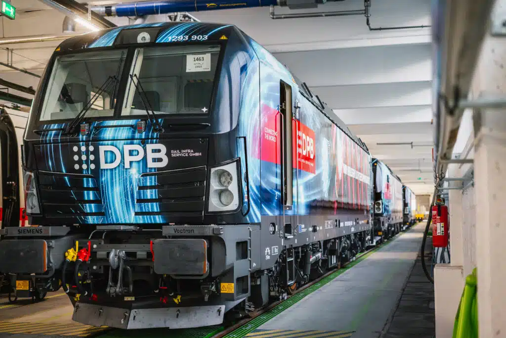 Eine moderne Elektrolokomotive des Typs Siemens Vectron steht in einer Wartungshalle. Die Lokomotive ist mit einer auffälligen blauen Folierung gestaltet, die ein dynamisches Lichtdesign zeigt, kombiniert mit dem Logo 'DPB Rail Infra Service GmbH'. Ein rotes Banner mit der Aufschrift 'Connecting the World' ist ebenfalls auf der Seite angebracht. Die Lok steht auf einem Gleis mit grünen Markierungen, umgeben von industriellen Strukturen wie Rohren, Kabeln und Feuerlöschern an der Wand. Die Perspektive des Bildes führt entlang der Lokomotive in die Tiefe der Halle.