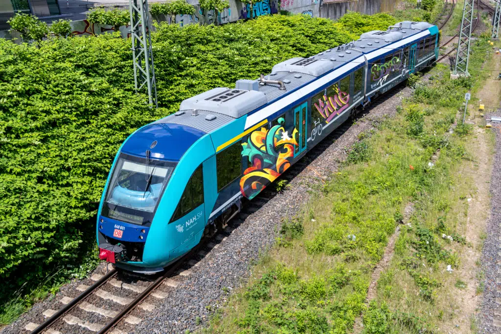 Ein moderner, blau-türkiser Regionalzug fährt auf einer eingleisigen Strecke durch eine grüne Umgebung. Die Seiten des Zuges sind mit farbenfrohen Graffiti-Motiven gestaltet, die geschwungene Muster und Schriftzüge enthalten. Der Blickwinkel von oben zeigt das Zugdach mit Lüftungseinheiten und technischen Vorrichtungen. Im Hintergrund sind eine mit Graffiti verzierte Betonwand, Oberleitungsmasten und dichte Vegetation zu sehen