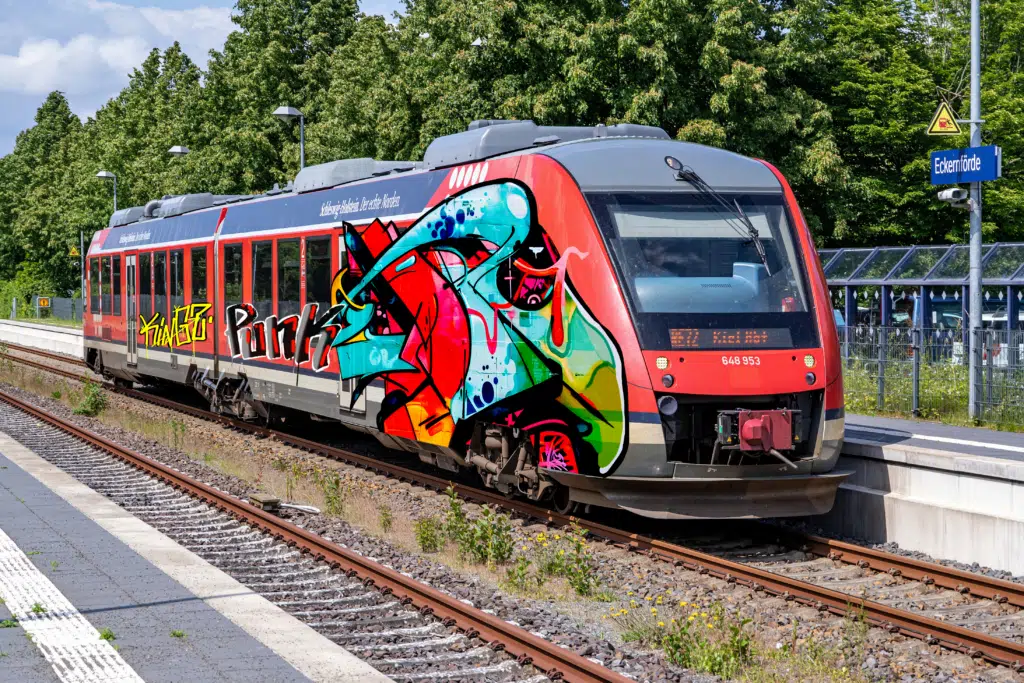 Ein roter Regionalzug steht am Bahnhof Eckernförde. Die Zugfront und die Seitenwand sind mit einem großflächigen, bunten Graffiti-Design versehen, das abstrakte Formen, Schriftzüge und leuchtende Farben kombiniert. Im Hintergrund sind Bäume, ein Wartehäuschen mit Fahrradständern sowie ein Stationsschild mit der Aufschrift 'Eckernförde' zu sehen. Der Himmel ist teilweise bewölkt, und das Sonnenlicht reflektiert auf der glänzenden Oberfläche des Zuges.