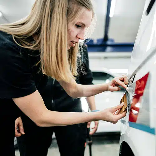 Eine junge Frau mit langen blonden Haaren arbeitet konzentriert an der Folierung eines Fahrzeugs. Sie trägt ein schwarzes T-Shirt und nutzt eine Rakel, um die Folie glatt auf die Karosserie aufzubringen. Im Hintergrund ist eine weitere Person in schwarzer Arbeitskleidung zu sehen, während mehrere weiße Fahrzeuge in einer gut beleuchteten Werkstatt stehen.